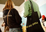 Two people are stood facing away from the camera, one person is wearing a late shows hoodie, the other is holding a late shows tote bag. 