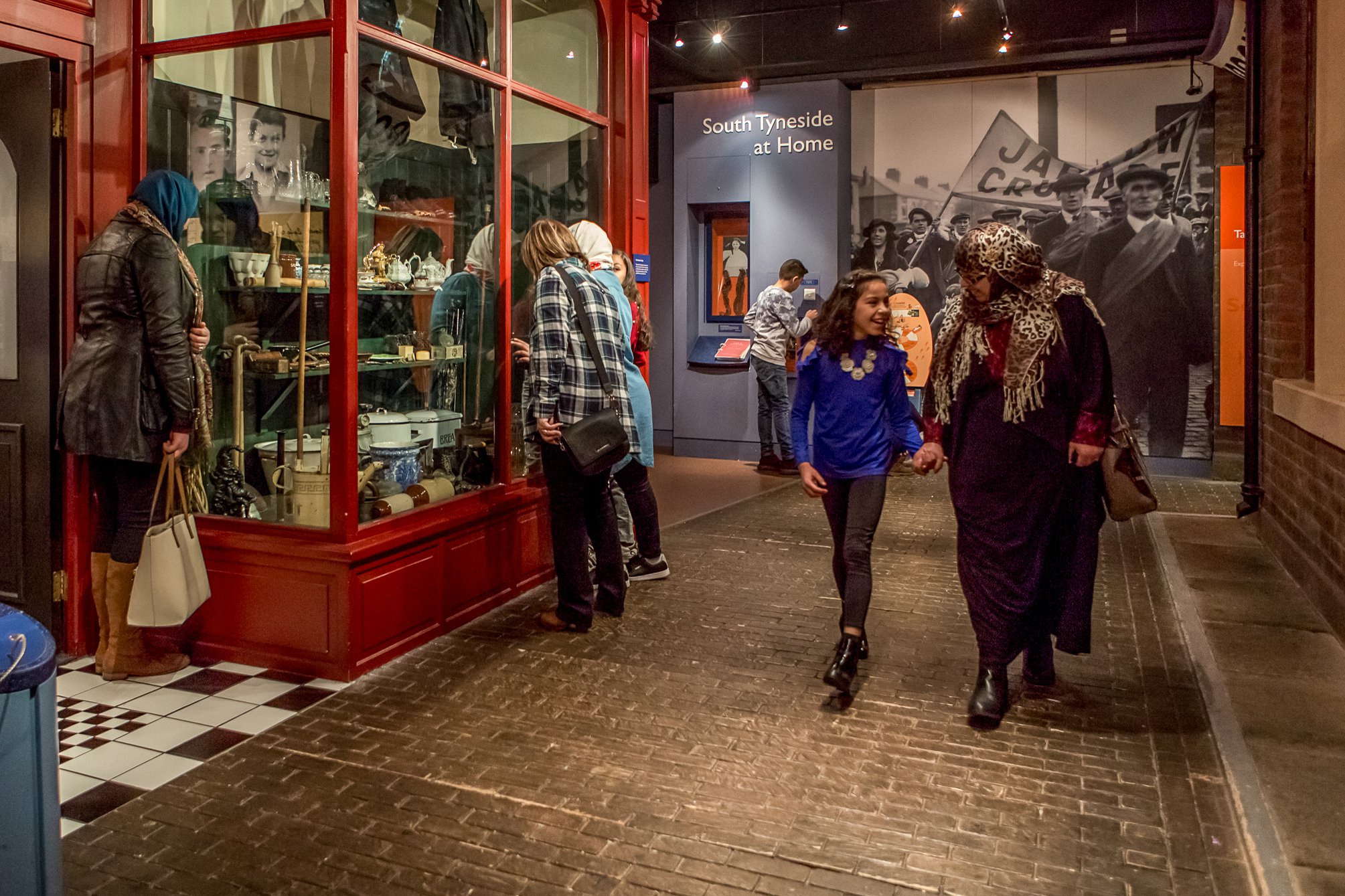 A busy museum exhibition which is designed as a street. Visitors are looking in shop windows, laughing and chatting
