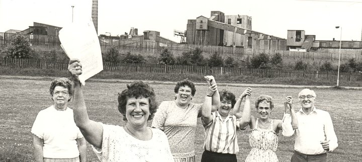 Black-and-white historical photograph of six community members standing on a grassy field in front of an industrial site. A brown haired woman in the foreground holds a document up.