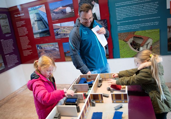 Three people, including two children and one adult, interact with a model display of the Commanding Officer's house in the reconstructed room. The background features informational panels about the fort