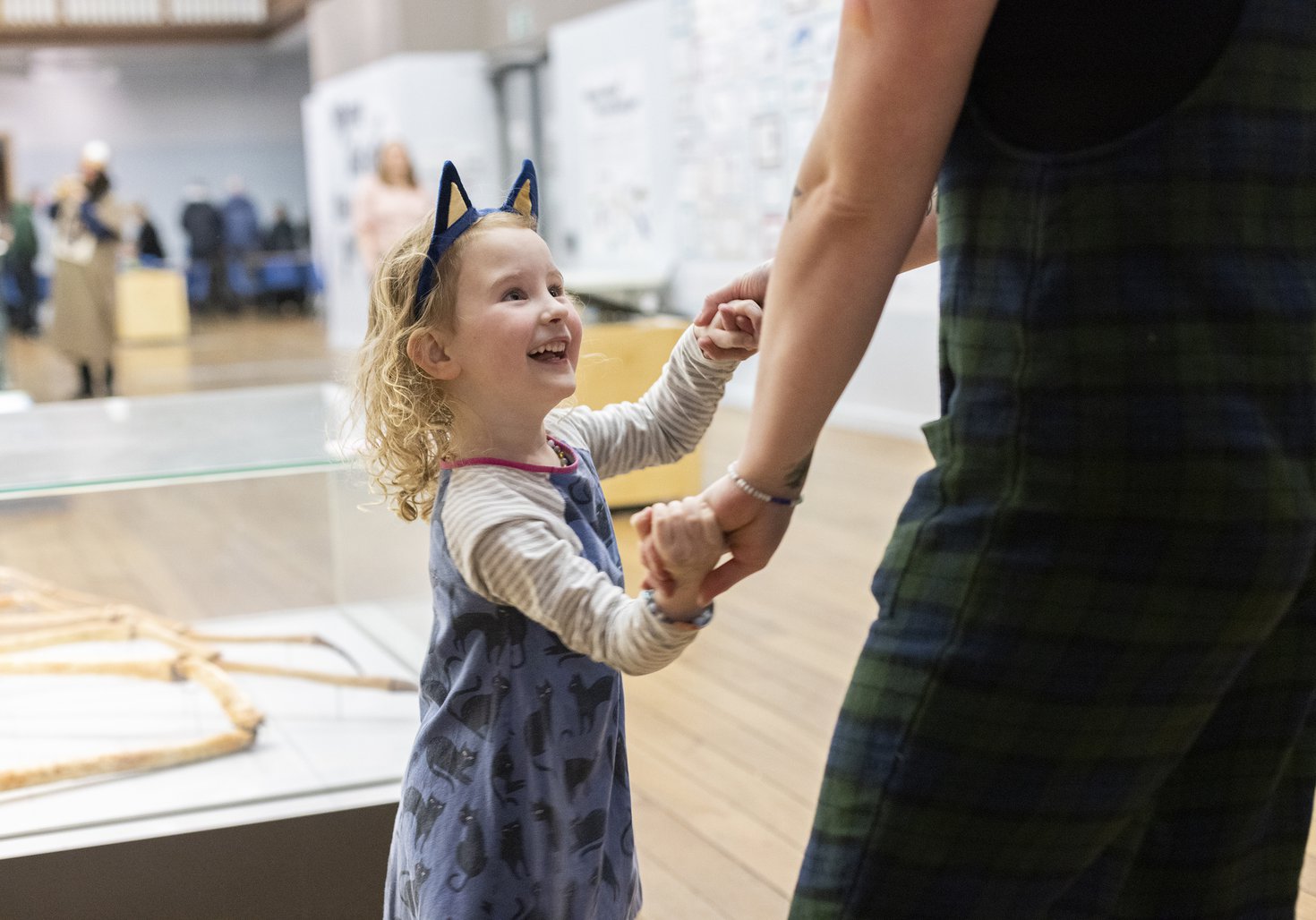 A child wearing cat ears stands in a museum. They are holding a parents' hand and looking up at them, smiling and laughing
