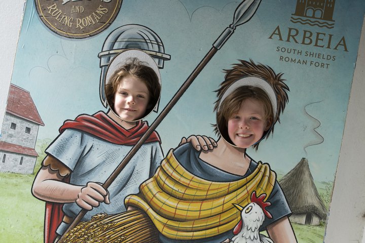 Two young children sticking their faces through cut out of Roman people