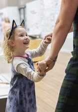 A child wearing cat ears stands in a museum. They are holding a parents' hand and looking up at them, smiling and laughing