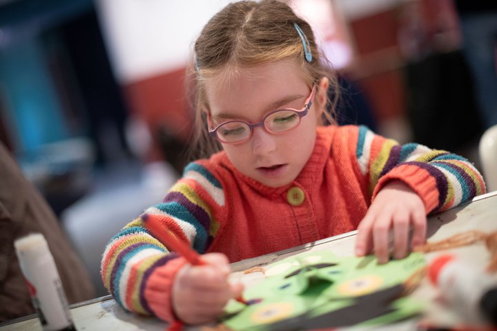 A photograph of a young girl colouring. She wears a striped jumper and glasses.