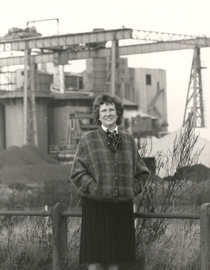 black and white photo of smiling woman with short brown curly hair standing in front of what appears to be an industrial site