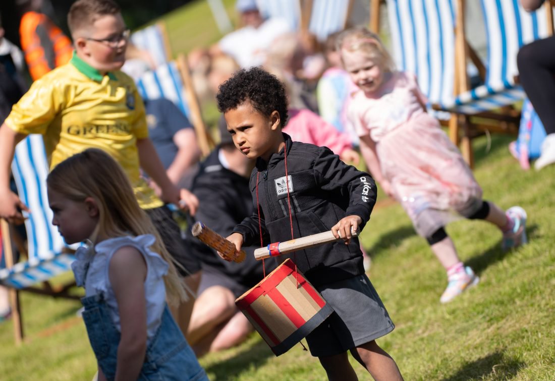 A boy wears a cardboard drum around his neck. He is surrounded by other children playing on grass. A boy wears a cardboard drum around his neck. He is surrounded by other children playing on grass.