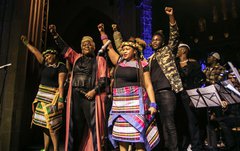 A group of people stand on a stage, they are smiling and each person has one arm raised in the air.