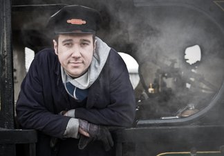 Man wearing railway cap with badge standing on the footplate of a steam engine