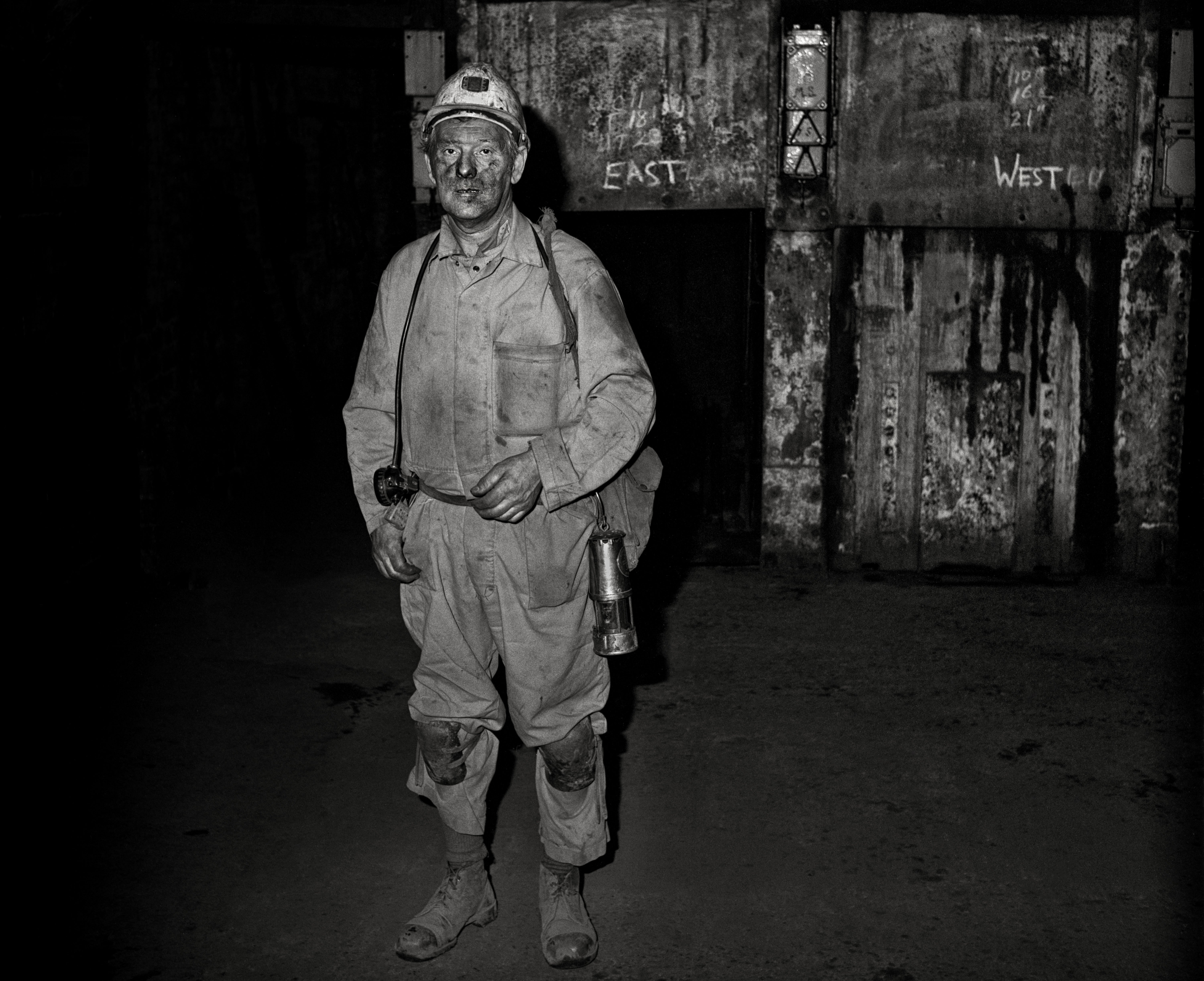 A miner stands in the Woodhorn Colliery pit. He is wearing overalls, a hard hat and carrying a torch. Behind him is the words 'East' and 'West'. A miner stands in the Woodhorn Colliery pit. He is wearing overalls, a hard hat and carrying a torch. Behind him is the words 'East' and 'West'.