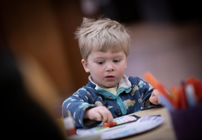A child with short blonde hair sitting at a table and taking part in a craft activity