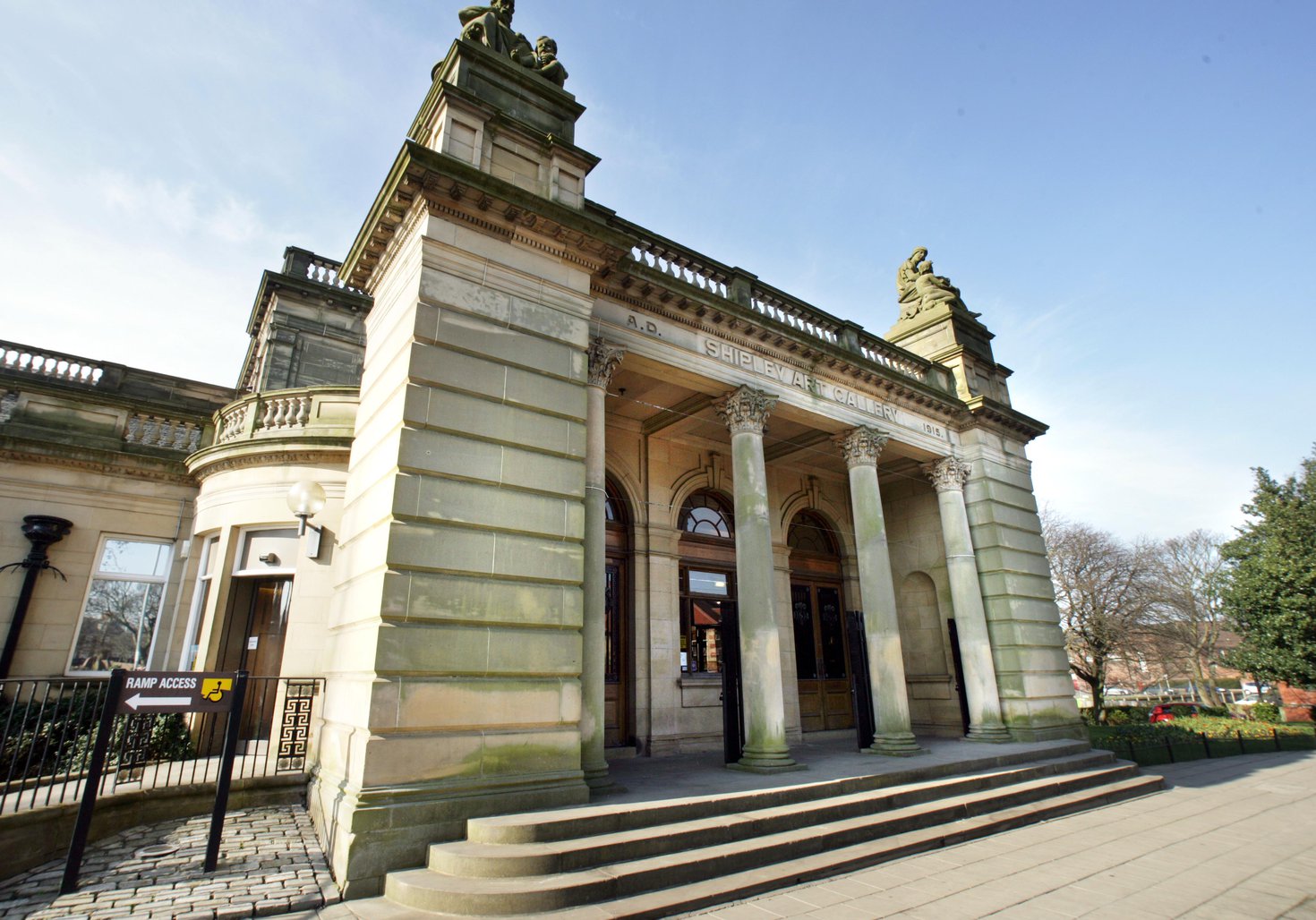 A photograph of the Shipley Art Gallery exterior. The building is made of yellow stone and the front has stone columns and two statues on the roof. 