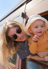 Woman with blonde hair and wearing sunglasses and her small child with mop hat leaning out of the window of a railway carriage