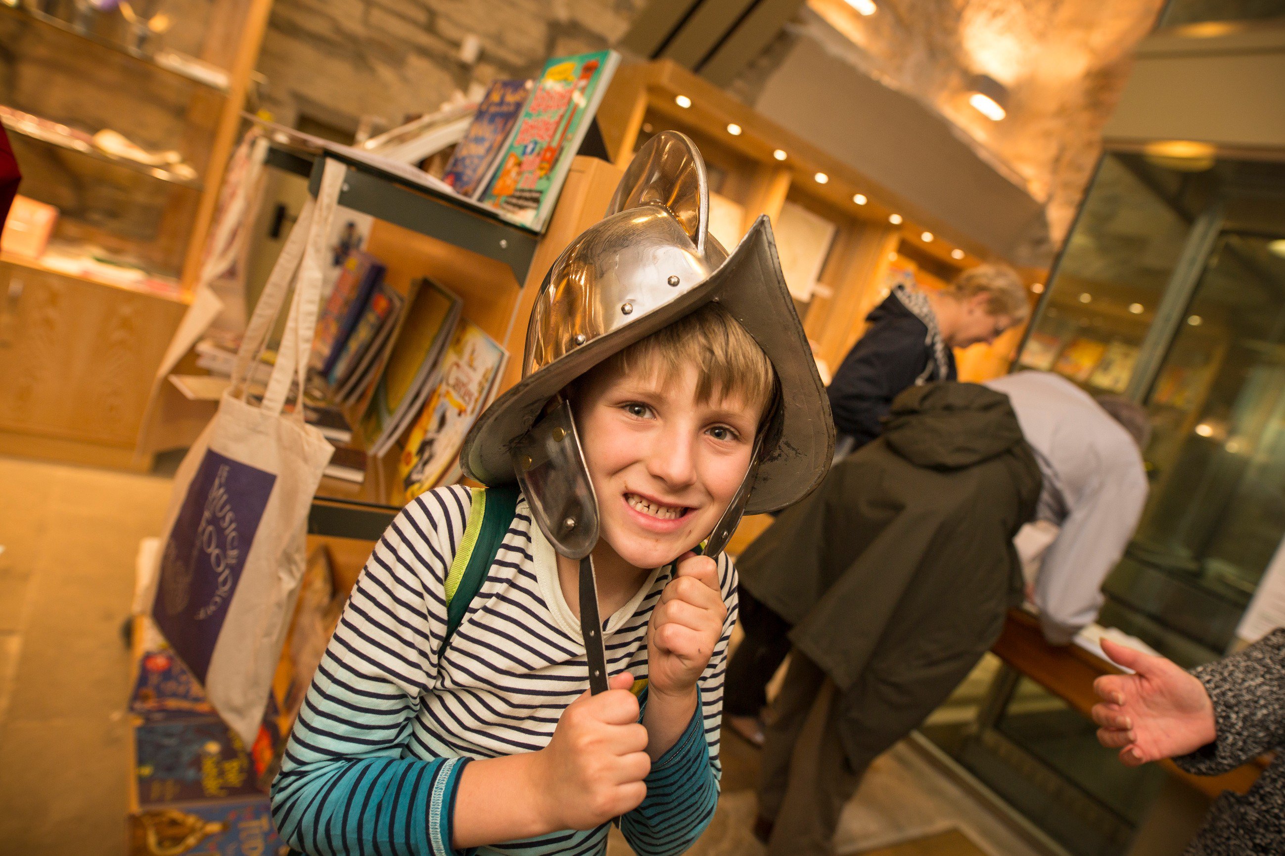 A child stands in a museum shop wearing a medieval helmet and smiling at the camera