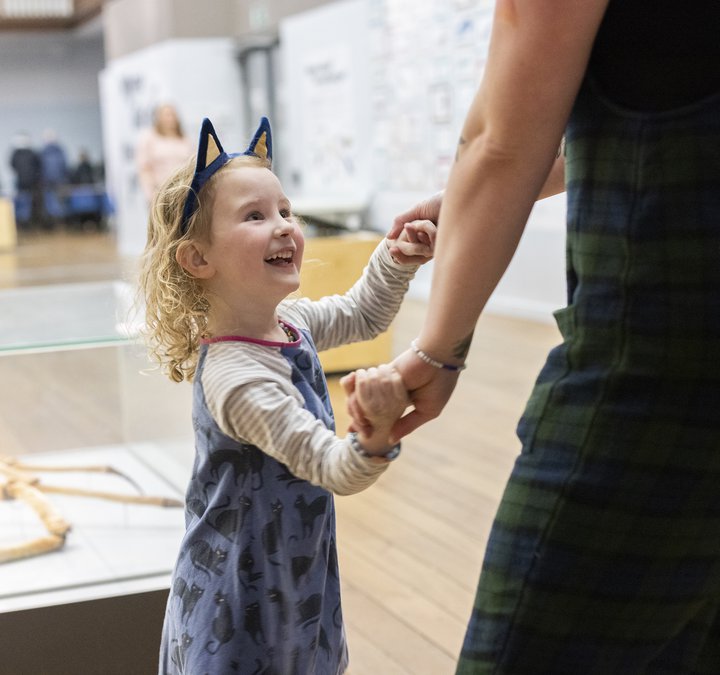 A small child wearing cat ears holds their parent's hand in a museum. She is smiling and laughing