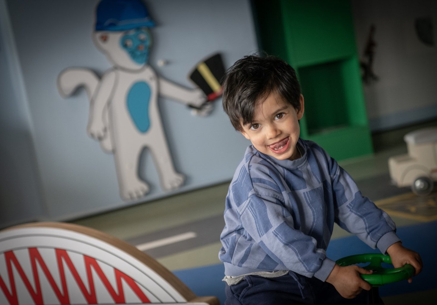 A small child looks at the camera, tongue sticking out. They are sat down, holding on to a toy, plastic steering wheel. In the background is a model of a bridge and an illustration of a cat holding a hat
