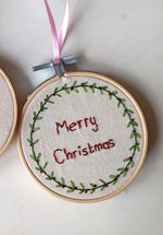 Two embroidery rings with cream fabric. One has a green and white mistletoe design and the other reads 'Merry Christmas' in red thread with a green wreath border