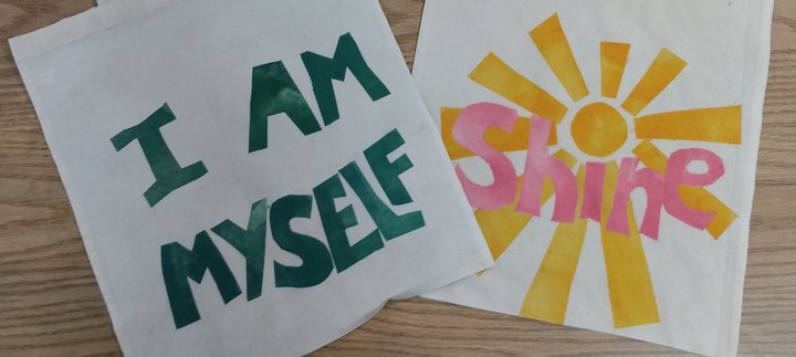 Two cotton tote bags laid on a table. One reads 'I am myself' in green text and the other 'Shine' in pink.