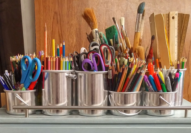 A photograph of different art materials in metal cups- pencils, paintbrushes, scissors, and more