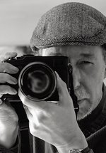 A portrait of a man wearing a flat cap, holding a camera and looking down the viewfinder. 