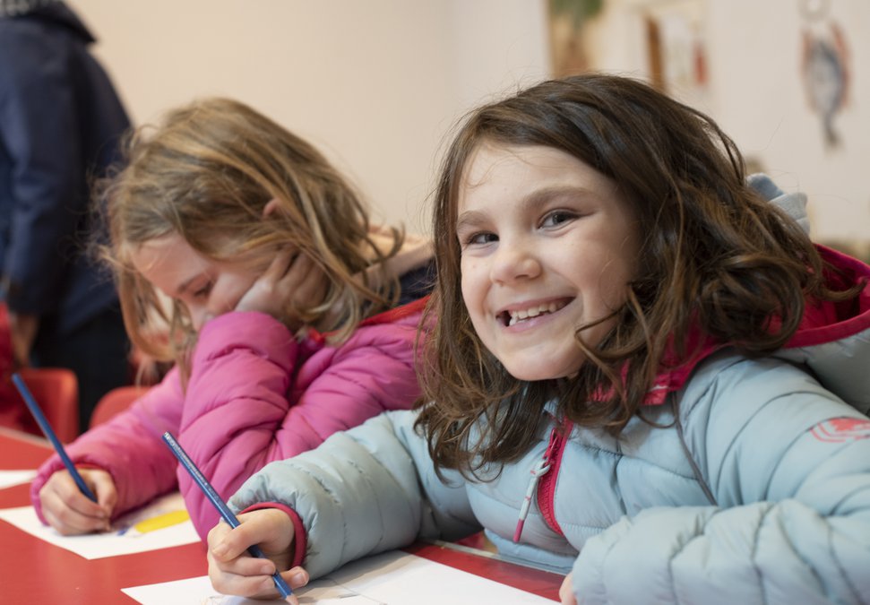 Two children sat at a table, one looks directly at the camera, grinning