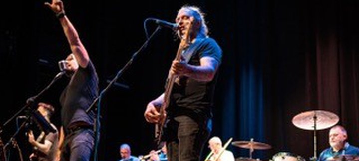 Band performing on stage with various instruments including guitar, drums, and brass.