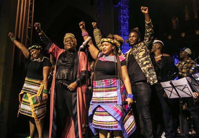 A group of people stand on a stage, they are smiling and each person has one arm raised in the air.
