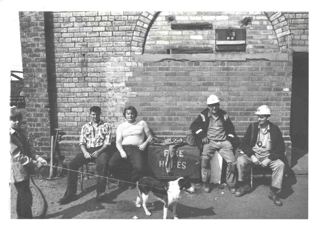 A black and white image of five men at Woodhorn Colliery sat down and having a cigarette break. One is stood up and holding the leash of a dog. Two men are wearing hard hats.
