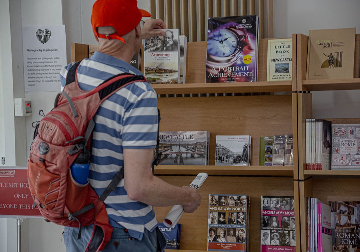 Man in red cap and blue and white striped t-shirt reaching to pick up a book from the shelves of books in Segedunum gift shop 
