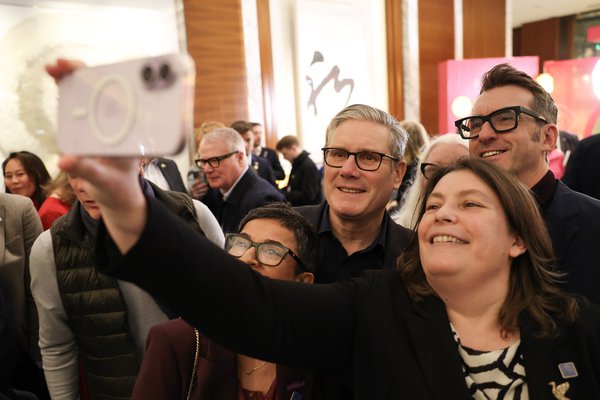 A woman holding a smartphone in front of her to take a photograph of the Prime Minister with herself, a man and a woman, in a room crowded with people. 