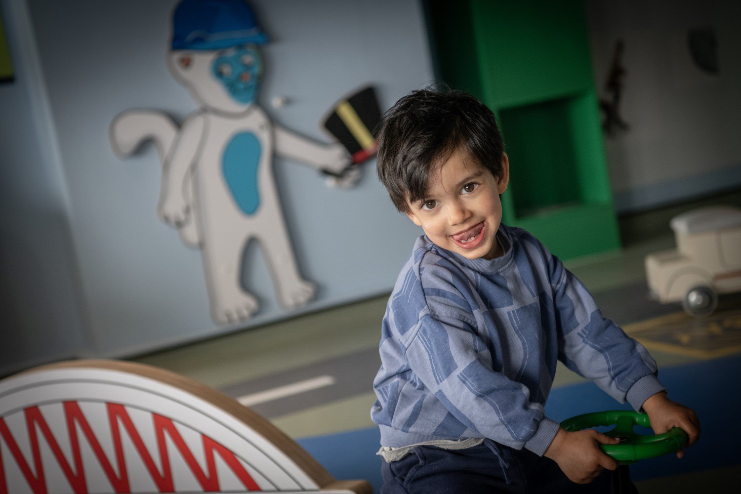 A small child looks at the camera with their tongue sticking out. They are sitting holding a toy, plastic steering wheel. In the background is a model bridge and an illustration of a cat holding a hat. A small child looks at the camera with their tongue sticking out. They are sitting holding a toy, plastic steering wheel. In the background is a model bridge and an illustration of a cat holding a hat.