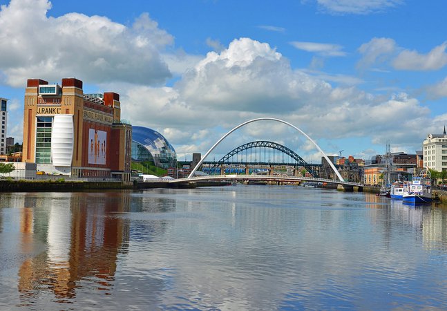 A large grey bridge and a large green bridge over a sunny river. To the left is a large red and yellow brick building which reads 'Baltic Flour Mills'.