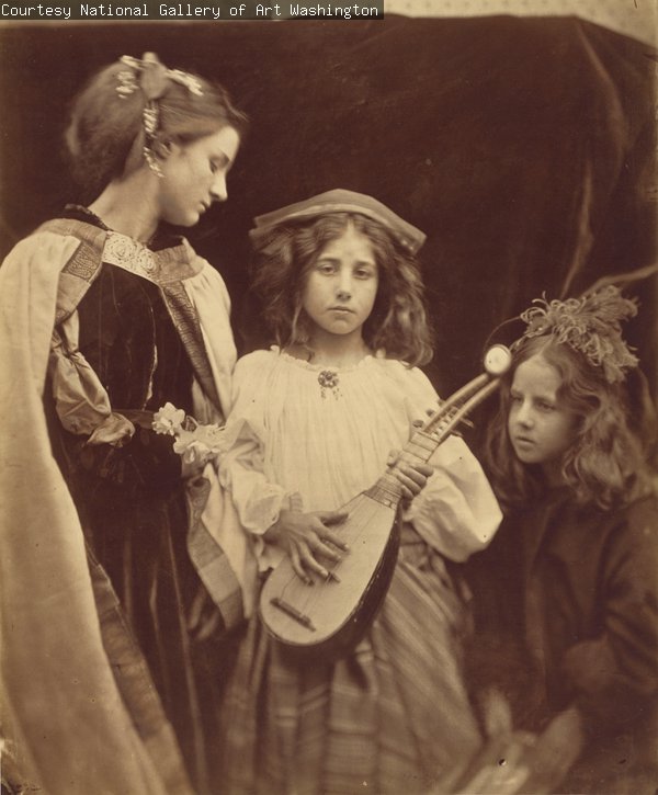 A sepia photograph of three children standing or seated in front of a dark background. They are wearing historical costumes. The child in the centre is holding a lute or similar instrument.