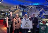 hotograph of 3 people in front of a T-Rex skeleton in the Fossil Stories Gallery at Great North Museum: Hancock being presented with a white board reading Autism Acceptance by another person.