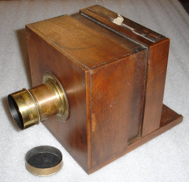 A cuboid shaped, varnished wood box camera with a cylindrical brass lens, on a white background.