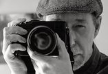 A close up portrait of a man wearing a hat and holding a camera up to his face 