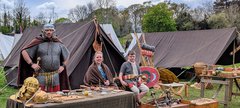 Historical reenactment camp with people in period costumes, tents, shields, helmets, and wooden tables with various items