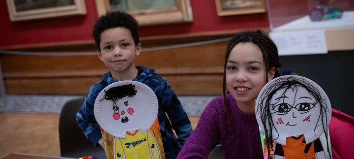 Two children smiling at the camera and holding up self portraits with paper plates.