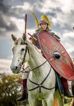 Two Roman soldiers on horseback, one with a spear and shield on a white horse, the other with a raised sword on a black horse, ready for battle under an overcast sky.