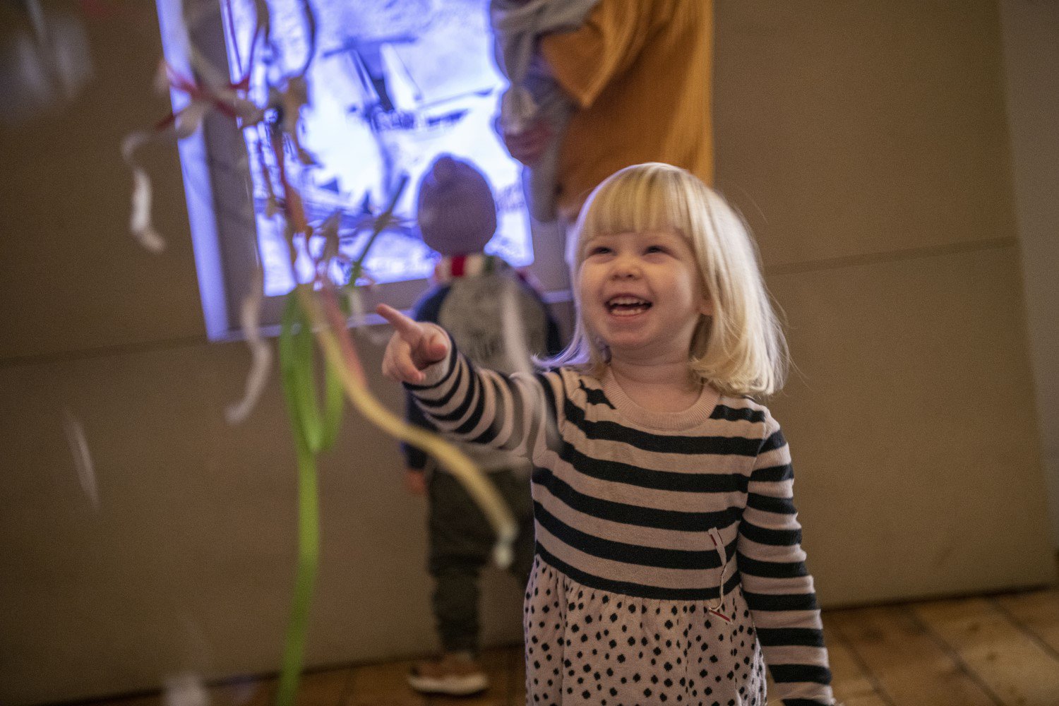 A small child is laughing and pointing at a museum display. In the background is a child watching a film projection on a wall. A small child is laughing and pointing at a museum display. In the background is a child watching a film projection on a wall.