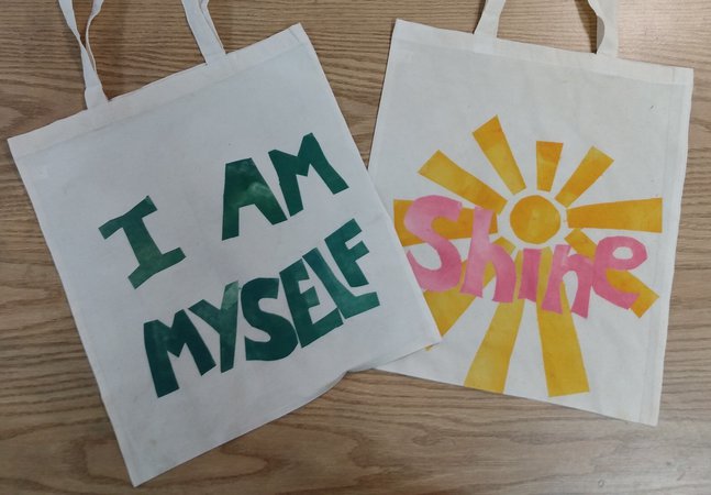 Two cotton tote bags laid on a table. One reads 'I am myself' in green text and the other 'Shine' in pink.