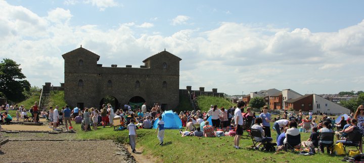People standing and sitting in front of a Roman style gatehouse on a sunny day