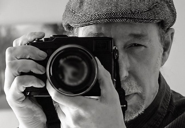 A portrait of a man wearing a flat cap, holding a camera and looking down the viewfinder. 