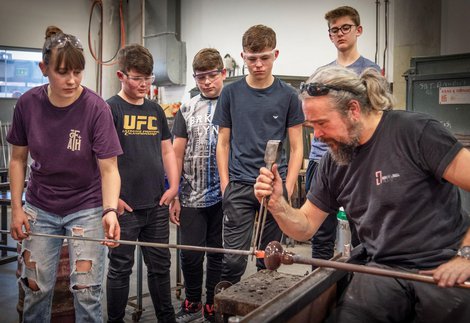 A group of students wearing eye protection gather round an artist doing glass blowing