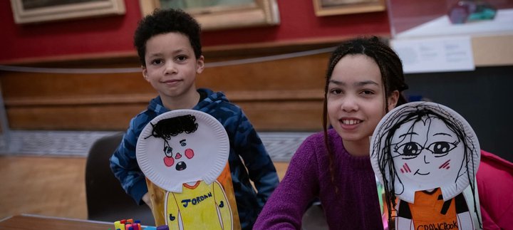 Two children smiling at the camera and holding up self portraits with paper plates.