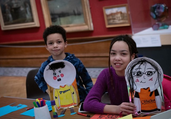Two children smiling at the camera and holding up self portraits with paper plates.