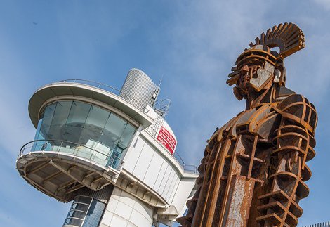 Rusted centurion sculpture with the tall glass viewing tower in the background against a blue sky