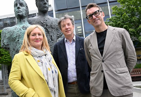 A woman and two men standing in front of sculpture of 2 figures and a tall building