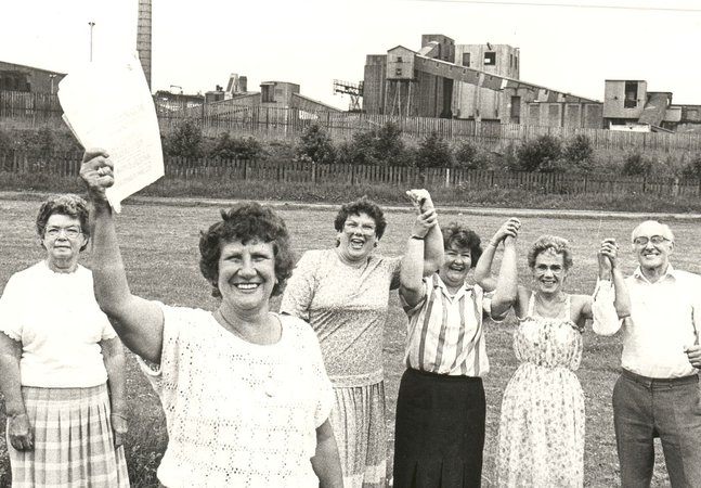 Black-and-white historical photograph of six community members standing on a grassy field in front of an industrial site. A brown haired woman in the foreground holds a document up.