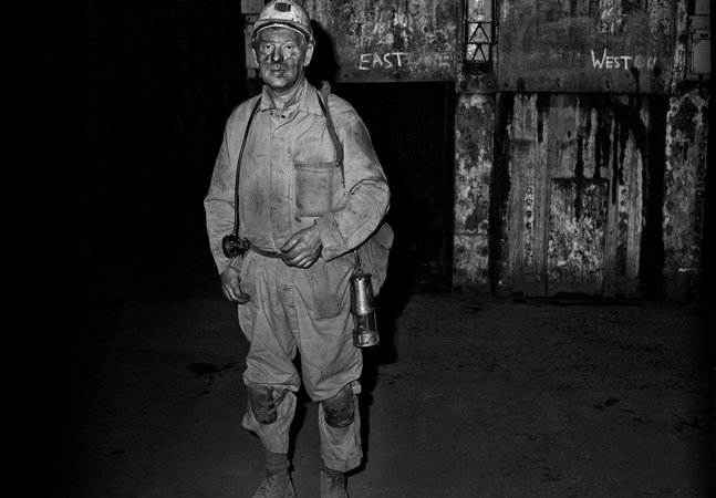 A miner stands in the Woodhorn Colliery pit. He is dressed in overalls, wearing a hard hat and carrying a torch. Behind him is the words 'East' and 'West' 