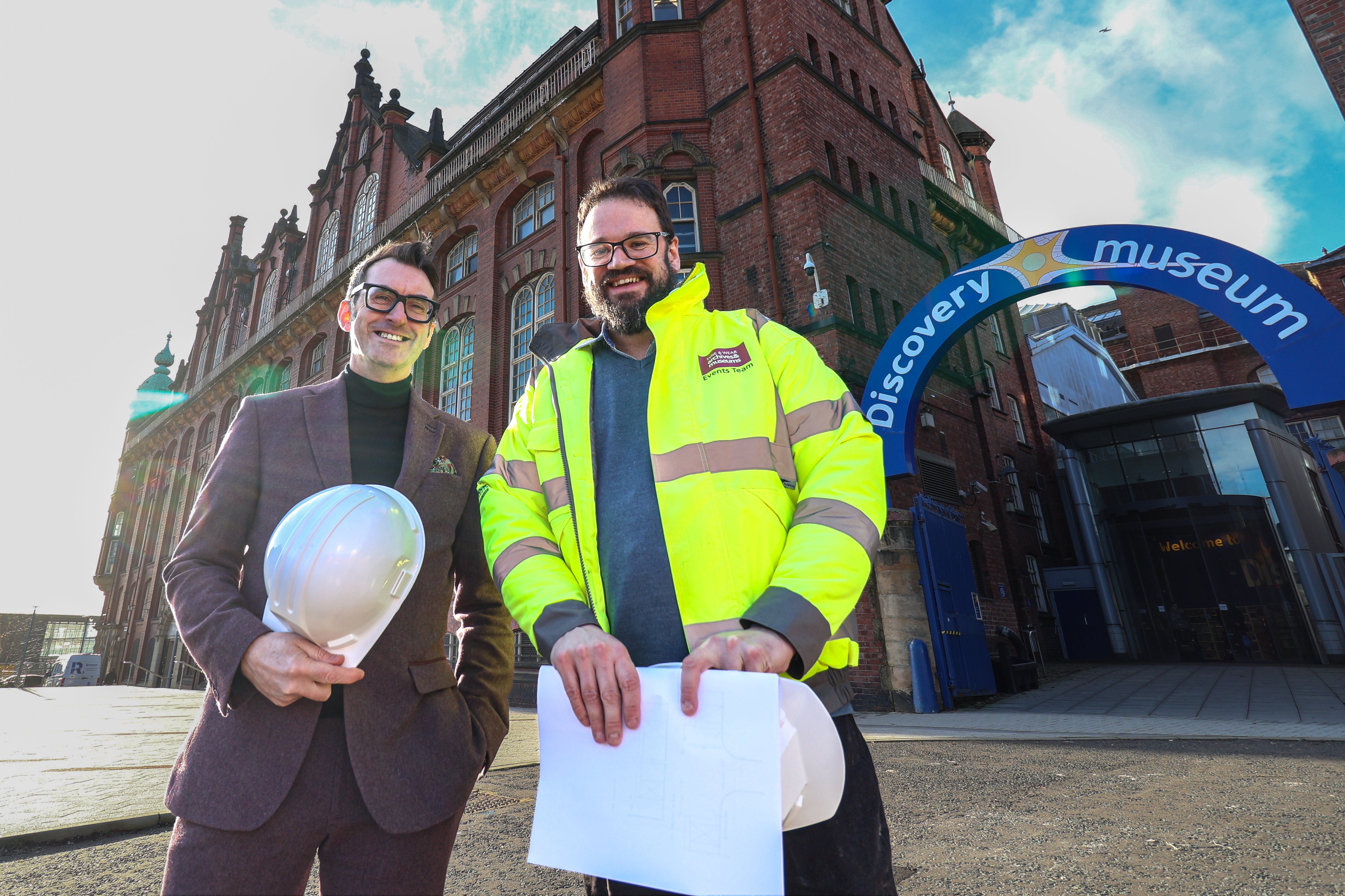 Two men smile outside Discovery Museum. One carries white hard hat, one wears high vis coat. Two men smile outside Discovery Museum. One carries white hard hat, one wears high vis coat.